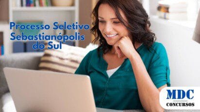 Mulher jovem de cabelos castanhos e ondulados, usando uma blusa verde, sorrindo enquanto olha para a tela de um laptop prateado em um ambiente aconchegante com sofá, almofadas e estante ao fundo - Sebastianópolis do Sul (SP) lança Processo Seletivo