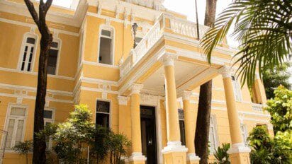 Fachada da sede da FUNCEB, um casarão histórico amarelo com detalhes brancos, colunas e vegetação ao redor.