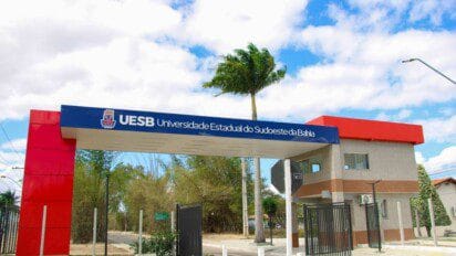 Entrada principal da Universidade Estadual do Sudoeste da Bahia (UESB) com portaria, portão aberto, placa azul com o nome da universidade, céu parcialmente nublado e palmeira ao fundo. Foto: Divulgação/Ascom-Uesb