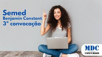 Mulher jovem de cabelos cacheados, usando blusa listrada e jeans, sentada no chão com as pernas cruzadas, segurando um laptop no colo. Ela sorri amplamente e levanta o dedo indicador da mão direita, como se tivesse uma ideia. O fundo é uma parede cinza lisa, e o chão tem textura de madeira - Semed Benjamin Constant (AM) convoca 86 aprovados em Processo Seletivo