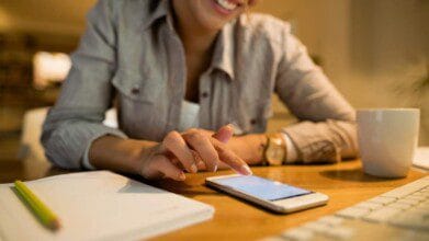 Mulher sorridente sentada em uma mesa de trabalho, utilizando um smartphone com o dedo indicador. Ela veste uma camisa listrada e relógio dourado no pulso esquerdo. Na mesa há um caderno branco com lápis, uma xícara branca e parte de um teclado de computador. O ambiente é bem iluminado e transmite sensação de conforto e concentração. (SENAI PI)