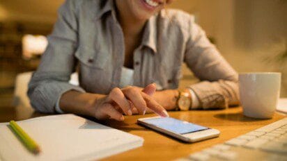 Mulher sorridente sentada em uma mesa de trabalho, utilizando um smartphone com o dedo indicador. Ela veste uma camisa listrada e relógio dourado no pulso esquerdo. Na mesa há um caderno branco com lápis, uma xícara branca e parte de um teclado de computador. O ambiente é bem iluminado e transmite sensação de conforto e concentração. (SENAI PI)