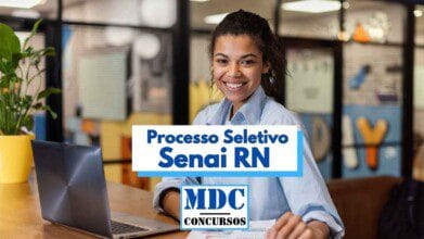 Mulher jovem sorridente está sentada à mesa em um ambiente moderno de escritório, com um notebook aberto à sua frente e documentos sobre a mesa. Ela usa camisa azul clara e transmite uma expressão amigável e confiante. Ao fundo, há divisórias de vidro com elementos coloridos e plantas decorativas. Em destaque na imagem, há os dizeres "Processo Seletivo Senai RN" e "MDC Concursos".