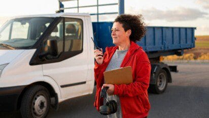 Mulher com rádio comunicador e prancheta em frente a caminhão branco e carreta azul. Foto: Freepik