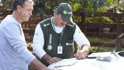 Técnico do SENAR RS preenche documentos ao ar livre enquanto conversa com produtor rural.