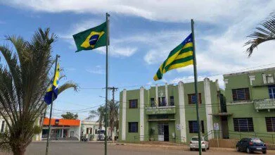 Imagem da fachada da Prefeitura de Silvânia-GO, com prédio verde e branco, letreiro visível, bandeiras hasteadas e cenário urbano com árvores, carros e banco ao fundo.