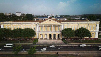 Fachada histórica do TJPA em Belém exibe arquitetura neoclássica em tons de amarelo e branco, com colunas, avenida movimentada em frente e contraste entre o prédio e o entorno urbano.