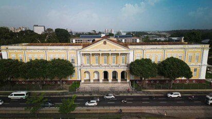 Fachada histórica do TJPA em Belém exibe arquitetura neoclássica em tons de amarelo e branco, com colunas, avenida movimentada em frente e contraste entre o prédio e o entorno urbano.