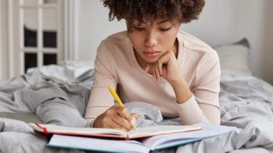 Jovem sentada em uma cama estudando e escrevendo em um caderno com uma caneta amarela.