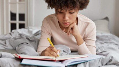 Jovem sentada em uma cama estudando e escrevendo em um caderno com uma caneta amarela.