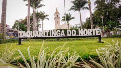A imagem mostra um letreiro com os dizeres “Taquaritinga do Norte” em letras brancas e grandes, instalado em uma área gramada bem cuidada, com plantas ornamentais na frente. Ao fundo, há uma igreja de arquitetura clássica com torre e cúpula dourada, cercada por palmeiras altas e outras árvores. O céu está nublado, e o ambiente transmite um ar tranquilo e acolhedor de cidade do interior.