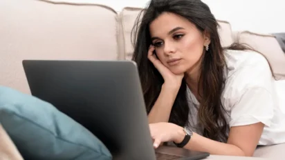 Mulher deitada no sofá olhando para a tela de um notebook com expressão pensativa. Foto: Freepik