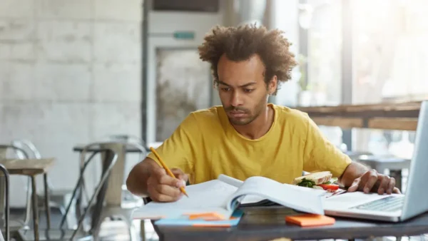 Terezinha/PE abre concurso com 49 vagas imediatas de até R$ 3.706,60 5 Homem sentado à mesa estudando com caderno aberto, lápis na mão e computador ao lado, em ambiente interno iluminado, com lanche sobre a mesa. Foto: wayhomestudio/Freepik