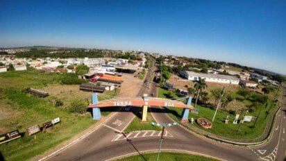 Vista aérea da entrada principal da cidade de Terra Boa, no Paraná, destacando um pórtico colorido com o nome "Terra Boa" em letras brancas. A imagem mostra ruas asfaltadas, comércios locais, áreas residenciais e vegetação ao redor. Em segundo plano, nota-se a expansão urbana da cidade, sob um céu azul claro e clima ensolarado, transmitindo um aspecto organizado e acolhedor do município.