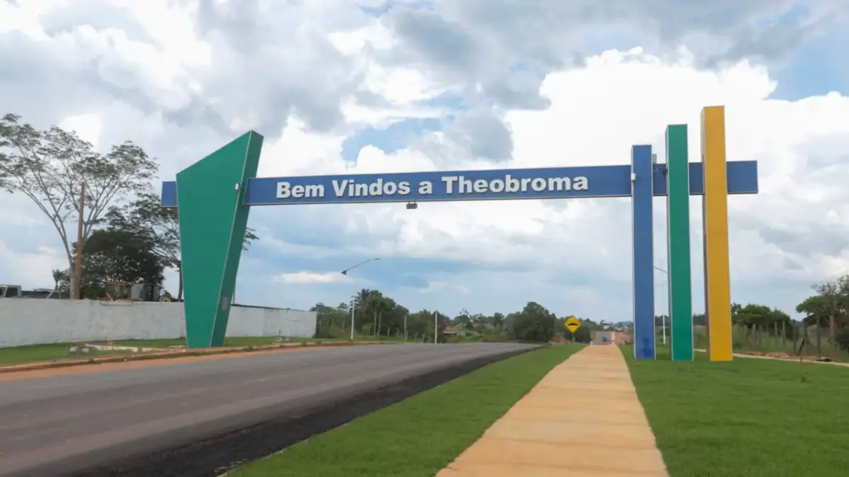 Theobroma/RO abre processo seletivo com salários de até R$ 5,2 mil 1 Portal na entrada da cidade de Theobroma, em Rondônia, com a frase Bem-vindos a Theobroma em uma estrutura azul sustentada por colunas coloridas em verde e amarelo sob um céu nublado.