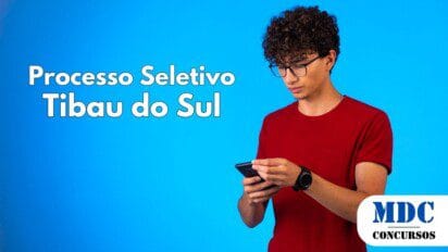 Jovem de cabelos cacheados, pele clara e óculos de grau, vestindo uma camiseta vermelha e um relógio preto, segurando e mexendo no celular com as duas mãos. O fundo é azul vibrante, criando um contraste com a roupa. Ele tem uma expressão concentrada enquanto olha para a tela do celular - Tibau do Sul (RN) oferece 13 vagas com salário de R$ 3 mil em Processo Seletivo