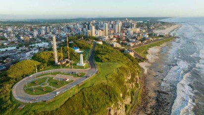 Vista aérea da cidade de Torres, no Rio Grande do Sul, mostrando falésias à beira-mar, área urbana com prédios altos e o litoral com ondas do oceano.