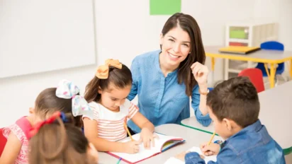 Professora sorridente sentada à mesa com crianças pequenas escrevendo em cadernos em sala de aula. Foto: tonodiaz/Freepik