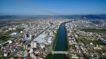 Vista aérea de Tubarão (SC) mostra área urbana densa, rio central com margens arborizadas, duas pontes, vias paralelas e montanhas ao fundo sob céu azul.
