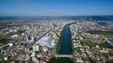 Processos Seletivos 6 Vista aérea de Tubarão (SC) mostra área urbana densa, rio central com margens arborizadas, duas pontes, vias paralelas e montanhas ao fundo sob céu azul.