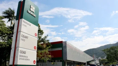 Totem de sinalização da UDESC em campus universitário, com prédios ao fundo, árvores e céu azul em Florianópolis. Foto: Divulgação/UDESC