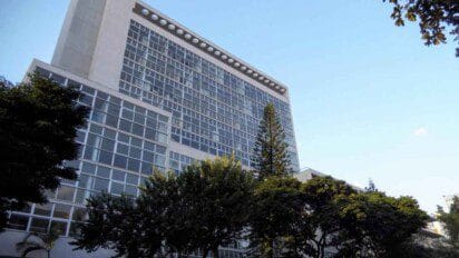 Prédio alto com fachada de vidro, cercado por árvores, sob céu claro e azul; imagem externa de uma unidade da Universidade do Estado de Minas Gerais.