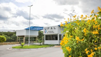Fachada da guarita de entrada do Campus do Sertão da UFAL, com estrutura branca e detalhes em azul. Ao lado, um arbusto com flores amarelas em destaque sob um céu nublado. Foto: Divulgação/UFAL