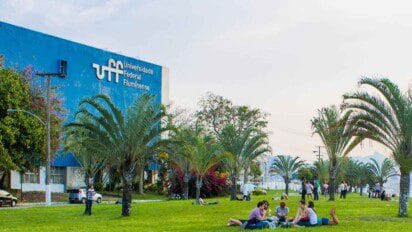 A imagem mostra o campus da Universidade Federal Fluminense (UFF), com destaque para um grande prédio azul à esquerda com o nome da instituição escrito em branco. Em frente ao prédio, há um extenso gramado com várias palmeiras e pessoas sentadas ou deitadas no chão, interagindo e aproveitando o ambiente ao ar livre. O céu está claro, e o clima parece agradável.