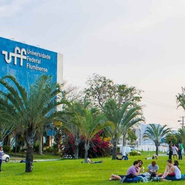 A imagem mostra o campus da Universidade Federal Fluminense (UFF), com destaque para um grande prédio azul à esquerda com o nome da instituição escrito em branco. Em frente ao prédio, há um extenso gramado com várias palmeiras e pessoas sentadas ou deitadas no chão, interagindo e aproveitando o ambiente ao ar livre. O céu está claro, e o clima parece agradável.