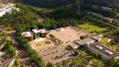Vista aérea do campus da UFJF com edifícios modernos, praça central ampla, áreas verdes e estacionamento ao redor.