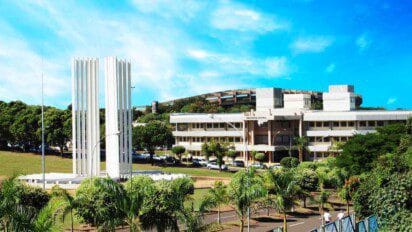 Vista do campus da UFMS com prédio da Biblioteca Central ao fundo, monumento branco em destaque e área arborizada sob céu azul.