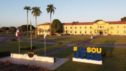 Fachada principal da UFRB com prédio amarelo, palmeiras, bandeiras hasteadas e letreiro colorido escrito #EUSOUUFRB. Foto: Divulgação/UFRB