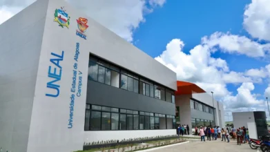 Fachada de prédio moderno da UNEAL Campus V, com letreiros azul e branco e brasão da universidade, sob céu azul com nuvens. No pátio de entrada, um grupo de pessoas aguarda e há motocicletas estacionadas.