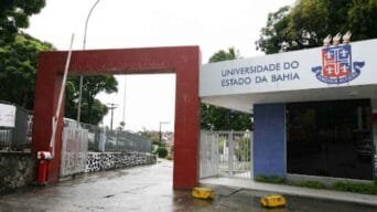 Portão de entrada da Universidade do Estado da Bahia com guarita, letreiro institucional e brasão da UNEB.