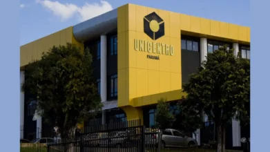 Fachada de prédio moderno com estrutura amarela e preta, exibindo o logotipo e o nome UNICENTRO Paraná em letras metálicas, cercada por árvores e uma grade frontal sob céu azul claro