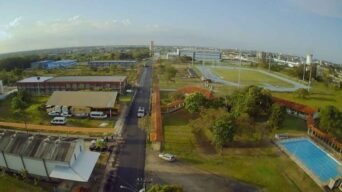 A imagem mostra uma vista aérea do campus da Universidade Federal do Amapá (UNIFAP) em Macapá. É possível ver diversas edificações distribuídas ao longo de uma avenida interna, com veículos estacionados ao lado de prédios administrativos e acadêmicos. À direita, há uma pista de atletismo cercada por gramado, além de uma piscina retangular e áreas arborizadas. Ao fundo, observa-se parte da cidade com construções urbanas e prédios. A imagem transmite um ambiente universitário estruturado, com áreas destinadas ao esporte e ao ensino.