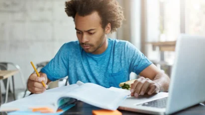 Homem estudando com livros e laptop enquanto segura um sanduíche em ambiente iluminado. Foto: wayhomestudio/Freepik