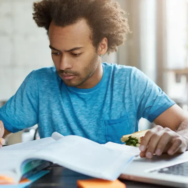 Homem estudando com livros e laptop enquanto segura um sanduíche em ambiente iluminado. Foto: wayhomestudio/Freepik