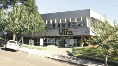 Fachada do prédio da UTFPR com o logotipo na entrada, cercada por árvores e com carro branco estacionado na via, campus que oferta especialização EaD gratuita