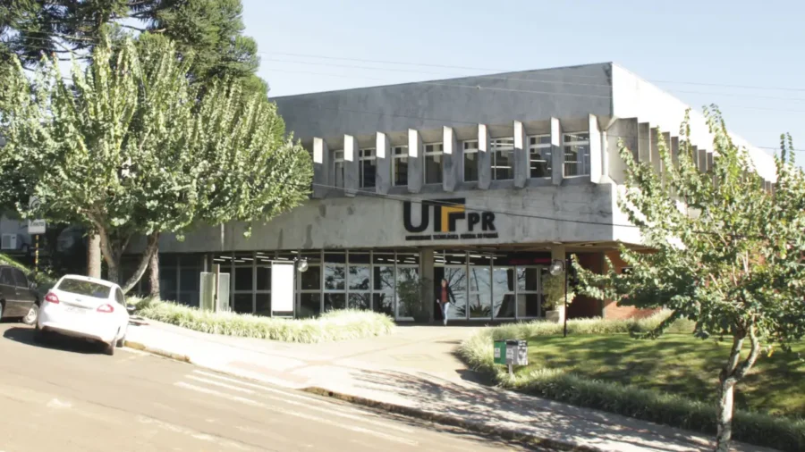 Fachada do prédio da UTFPR com o logotipo na entrada, cercada por árvores e com carro branco estacionado na via, campus que oferta especialização EaD gratuita