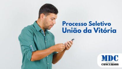 Homem de pele clara e barba curta, vestindo camisa verde de mangas dobradas, segurando e interagindo com um smartphone. Ele está concentrado na tela do dispositivo, com expressão séria. O fundo é um cenário simples e branco - União da Vitória (PR) oferece 60 Vagas