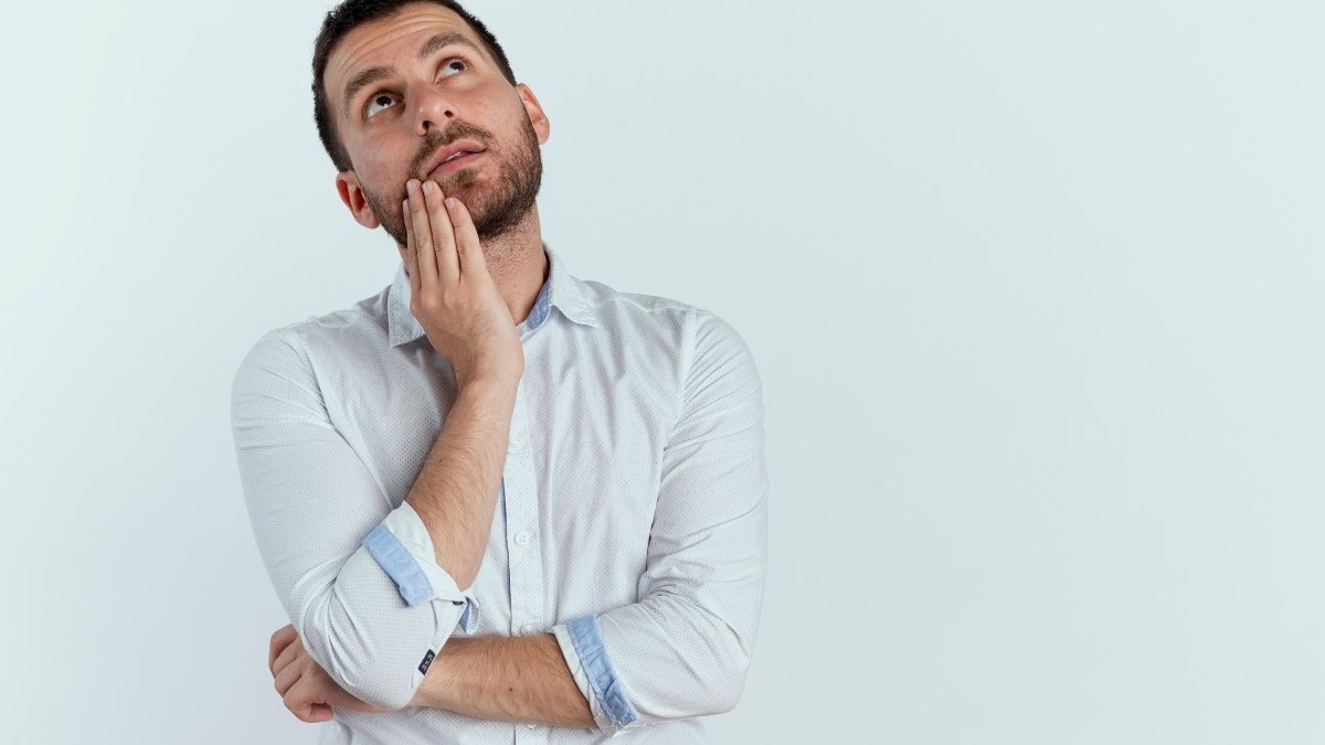Diferença entre concurso e processo seletivo: entenda antes de se inscrever 2 Homem de camisa social clara com expressão pensativa, olhando para cima com a mão no rosto, em frente a um fundo branco. Foto: stockking/Freepik