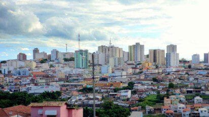 Vista panorâmica da cidade de Varginha, com prédios altos ao fundo e casas coloridas em primeiro plano sob céu parcialmente nublado.