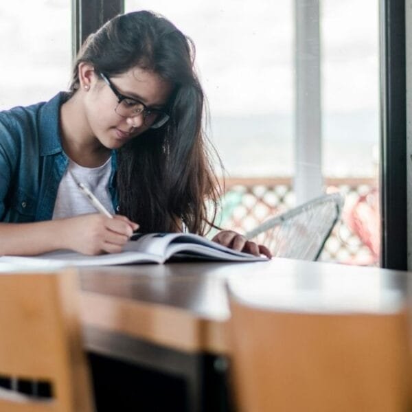 Estudante de óculos escrevendo em caderno aberto, sentada sozinha em mesa de biblioteca ou sala de estudos com vista externa. Foto: Kyle Gregory Devaras/Unsplash