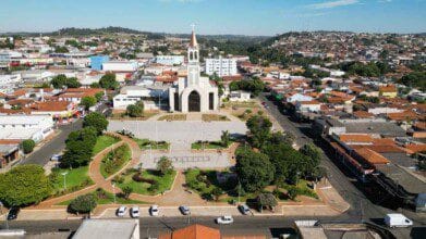 Processos Seletivos 2 Vista aérea do centro de Nerópolis, em Goiás, destacando uma ampla praça arborizada com caminhos pavimentados e jardins bem cuidados. Ao fundo, sobressai uma igreja de arquitetura imponente com torre e cruz no topo, localizada no coração da cidade. Ao redor, é possível observar um aglomerado urbano com casas, comércios e ruas asfaltadas, além de áreas mais elevadas com moradias ao longe.
