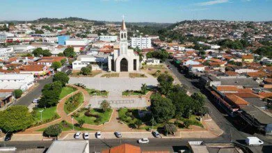 Vista aérea do centro de Nerópolis, em Goiás, destacando uma ampla praça arborizada com caminhos pavimentados e jardins bem cuidados. Ao fundo, sobressai uma igreja de arquitetura imponente com torre e cruz no topo, localizada no coração da cidade. Ao redor, é possível observar um aglomerado urbano com casas, comércios e ruas asfaltadas, além de áreas mais elevadas com moradias ao longe.