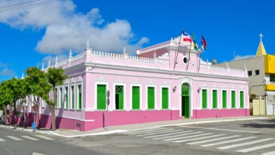 Prédio histórico da Prefeitura de Vitória da Conquista, com fachada rosa, portas e janelas verdes, bandeiras hasteadas e céu azul ao fundo em esquina urbana. Foto: Divulgação/Prefeitura de Vitória da Conquista-BA