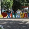 Letreiro turístico colorido com a frase "Eu amo Xinguara", onde a palavra "amo" é substituída por um coração vermelho, localizado em uma praça arborizada com parquinho infantil ao fundo.