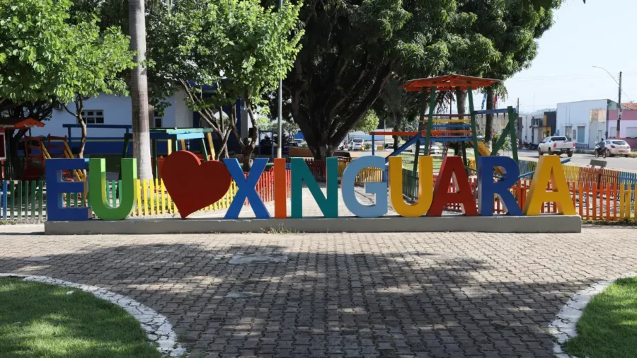 Letreiro turístico colorido com a frase "Eu amo Xinguara", onde a palavra "amo" é substituída por um coração vermelho, localizado em uma praça arborizada com parquinho infantil ao fundo.