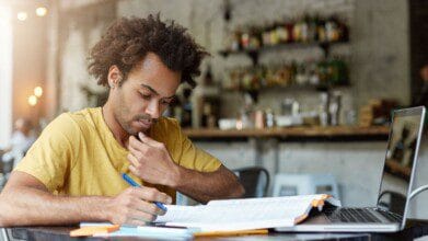 Homem jovem estudando em um local com laptop, livro aberto e anotações sobre a mesa. Foto: wayhomestudio/Freepik